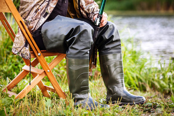 man sits in military uniform and rubber boots on a chair and catches fish on the river bank