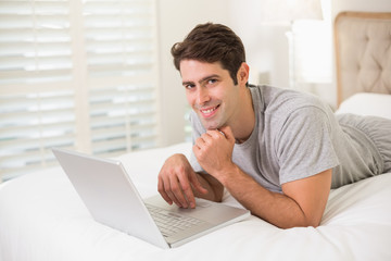 Casual smiling young man using laptop in bed