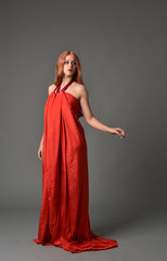 full length portrait of woman wearing red silk gown, on grey studio background.