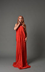 full length portrait of woman wearing red silk gown, on grey studio background.