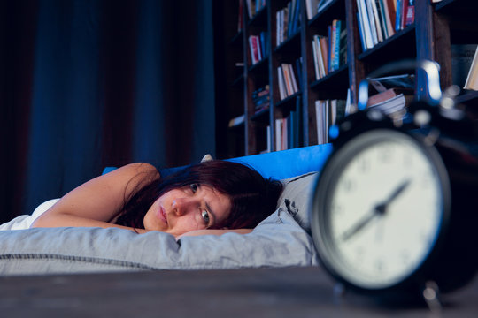 Photo of brunette with insomnia lying on bed next to alarm clock at night