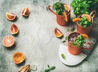 Blood orange Moscow mule alcohol cocktails with fresh mint leaves and friuts and ice in copper mugs on board over grey concrete background, copy space