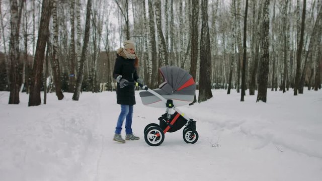 Side view of a young mother walking with a baby stroller in winter park