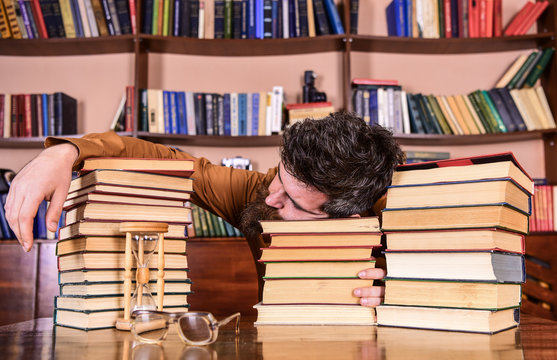 Overstudied concept. Man on sleeping face lay between piles of books, fall asleep while studying in library, bookshelves on background. Teacher or student with beard fall asleep on books, defocused.