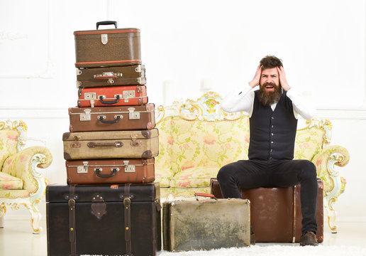 Man, butler with beard and mustache delivers luggage, luxury white interior background. Luggage and relocation concept. Macho elegant on painful face sits shocked near pile of vintage suitcase.