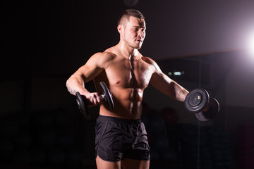 Fototapeta na wymiar Powerful bodybuilder doing the exercises with dumbbells. Strong male with naked torso on dark background. Strength and motivation.