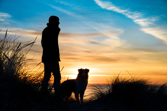 Silhouette von einem Mann und einen Hund am Strand bei Sonnenuntergang umrahmt von Gräsern.
