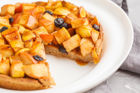 Whole vegan apple pie with cinnamon, raisins and caramel on white plate, gray background.