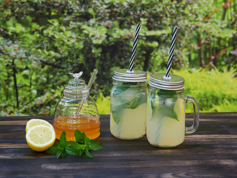 Two jars of fresh lemonade with sparkling water, mint and honey, set on a dark oak table, with green garden vegetation in the background.