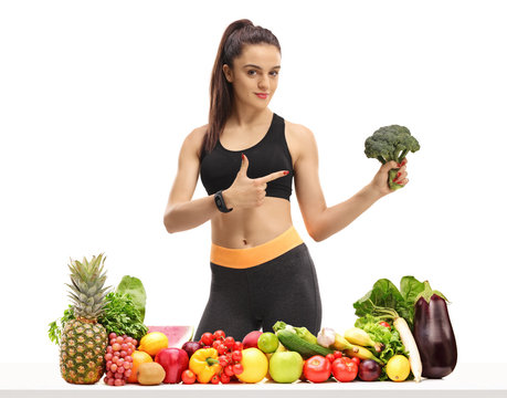 Fitness woman behind a table with fruit and vegetables holding broccoli and pointing