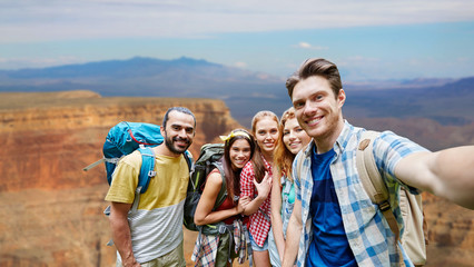 travel, tourism and technology concept - group of smiling friends or travelers with backpacks taking selfie over rocks of grand canyon national park background