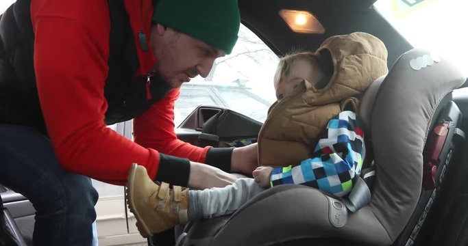 Father puts baby boy into protective seat and fastens straps in interior of car