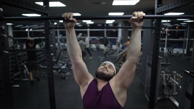 Overweight man trying to pull up on sport bar, weak body muscles, gym training 