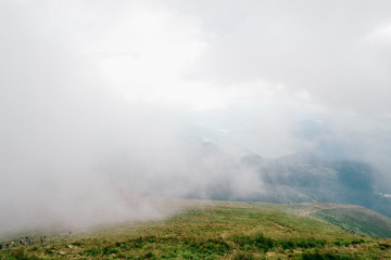 Foggy weather in mountains. Travelers hiking over misty hiils. Tourists climbing up. Healthy  activity. Summer moody scenic view from high altitude at nature outdoor. Beauftiful landscape. Trekking.