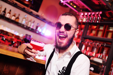young stylish cute guy in round sunglasses with cocktail having fun on bar background