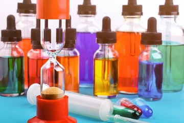 Closeup of hourglass on blue surface and background with glass bottles filled colored liquid and syringe