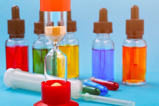 Closeup of hourglass on blue background with glass bottles filled colored liquid and syringe
