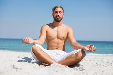 Fototapeta na wymiar Full length of shirtless man meditating at beach
