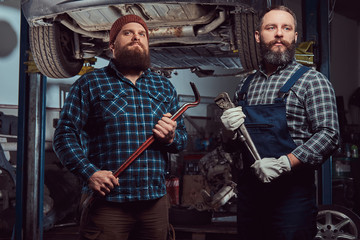 Two bearded brutal mechanics repair a car on a lift in a garage.