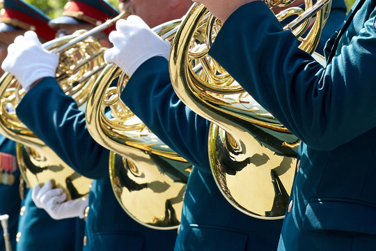 Closeup of military musicians in green uniform and white gloves playing a French horn during a parade in the street.