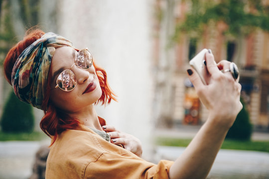 beautiful young woman wearing round sunglasses taking a selfie picture outdoors. hippie fashion blogger on vacation, taking self portrait with a smartphone. street style, music festival portrait.