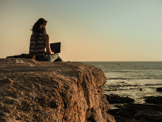 back of a sitting digital nomad woman on a rock at the beach working with her laptop during sunset