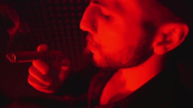 A young man smokes a cigar and releases smoke rings in a dark room