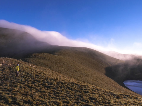 An asian man standing on the hill of mountains, and there is a thick fog drifting torward where he is.