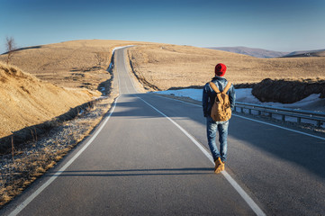 Fototapeta premium A bearded hipster with an old-fashioned vintage backpack wearing sunglasses with a red hat and jeans jacket and jeans walking goes with his back along a countryside asphalt mountain road in the