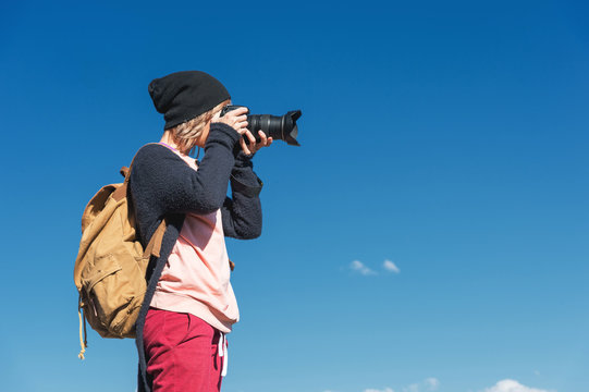 Portrait of a stylish girl hippy in a hat and with a backpack that takes pictures of her on a DSLR camera outdoors against a blue sky on a sunny afternoon. The concept of photography in travel