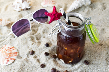 Cold coffee with ice in a bottle in the sand near the seashells and sunglasses. Summer concept