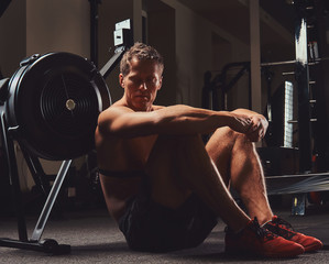 Muscular shirtless athlete sits on a floor resting after a hard workout in the gym.