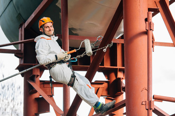 Industrial climber in helmet and uniform painting water tower. Professional Painter working on height. Risky job. Extreme occupation.