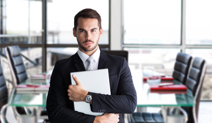 Businessman in suit posing with his laptop  against empty corporate meeting room 