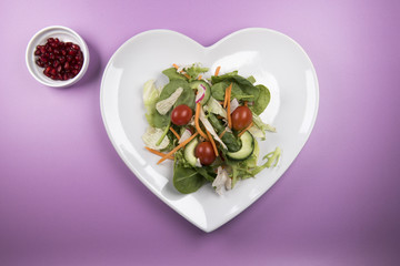 Traditional salad on a heart shaped plate with a bowl of pomegranate on a pink background