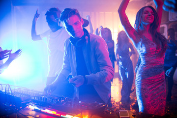 Waist up portrait of handsome young DJ standing at mixer making music during night party in club, scene lit by stage lights with beautiful girls wearing dresses dancing in background