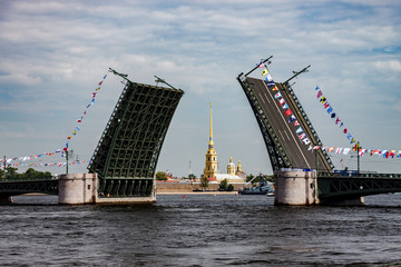 Fototapeta na wymiar A diluted palace bridge decorated with flags on the Neva River in the city of St. Petersburg. Between the diverged spans of the bridge is seen Peter and Paul Cathedral and the fortress. 