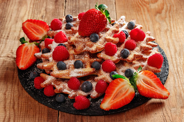 On a stone dish lie wafers sprinkled with powdered sugar, blueberries, raspberries and strawberry slices. A black dish of slate lies on a table of old wooden boards.