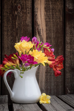 A bunch of colorful freesia flowers in a white ceramic pitcher on a rustic wooden plank table.