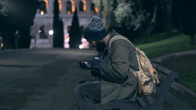 Lonely homeless sit on bench at night, cheking his alms