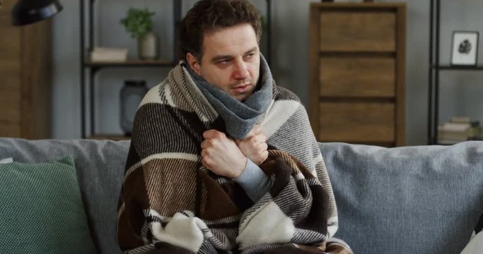Caucasian young ill man in the warm scarf shivering under the plaid on the couch in the living room as he having a high temperature during a flu. At home. Indoor
