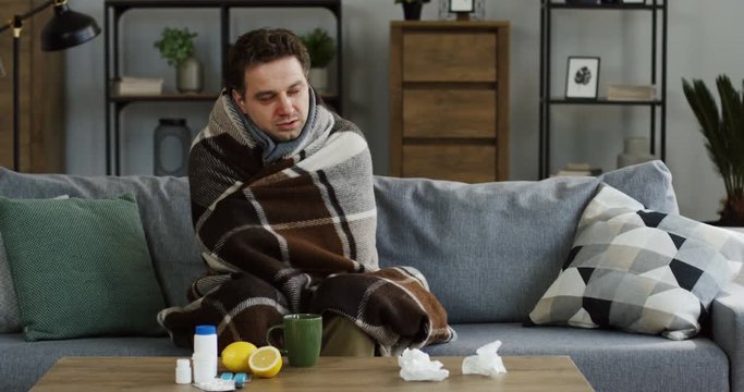 Young unwell caucasian man in the warm scarf shivering as he having a high temperature and a cold, sitting on the couch in the living room. Indoors