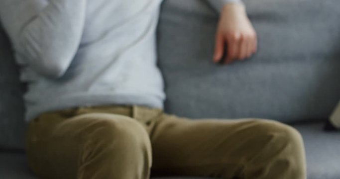 Close up of the young man in a gray scarf and a plaid talking on the phone while sitting on the couch, sneezing and caughing as having a flu. Indoor