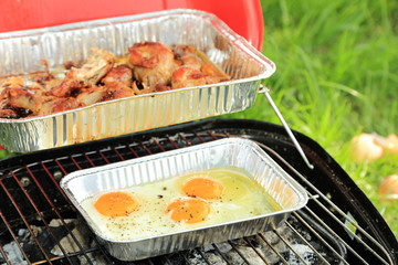 Smażenie jajek sadzonych i pieczenie udek z kurczaka na grilu turystycznym. - obrazy, fototapety, plakaty