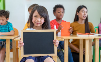 Kid girl holding blank blackboard with diversity friends and teacher at background,Kindergarten...