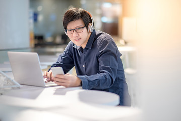 Young Asian business man listening to music by headphones and smartphone while working with laptop computer in co working space. freelance or digital nomad lifestyle concepts