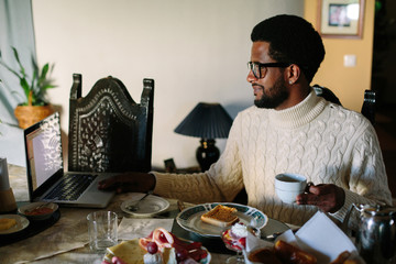 Portrait of young African man wearing glasses using laptop and have breakfast at home