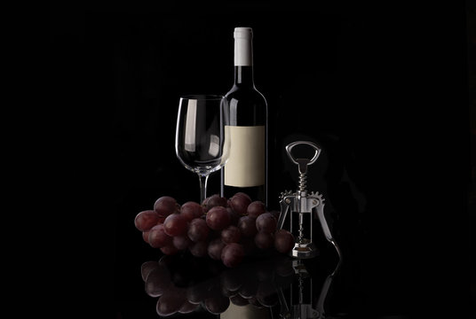 Red wine bottle, empty wine glass, corkscrew and grape on black background
