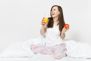 Young brunette woman sits on bed with white sheet, pillow, wrapping in blanket isolated on white background. Beauty female have breakfast with apple, fresh orange juice. Rest, relax, good mood concept