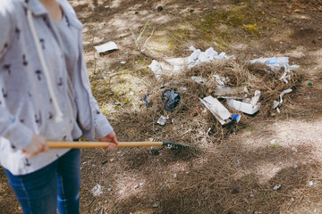 Cropped Young woman in casual clothes cleaning rubbish using rake for garbage collection in littered park. Problem of environmental pollution. Stop nature garbage, environment protection concept.